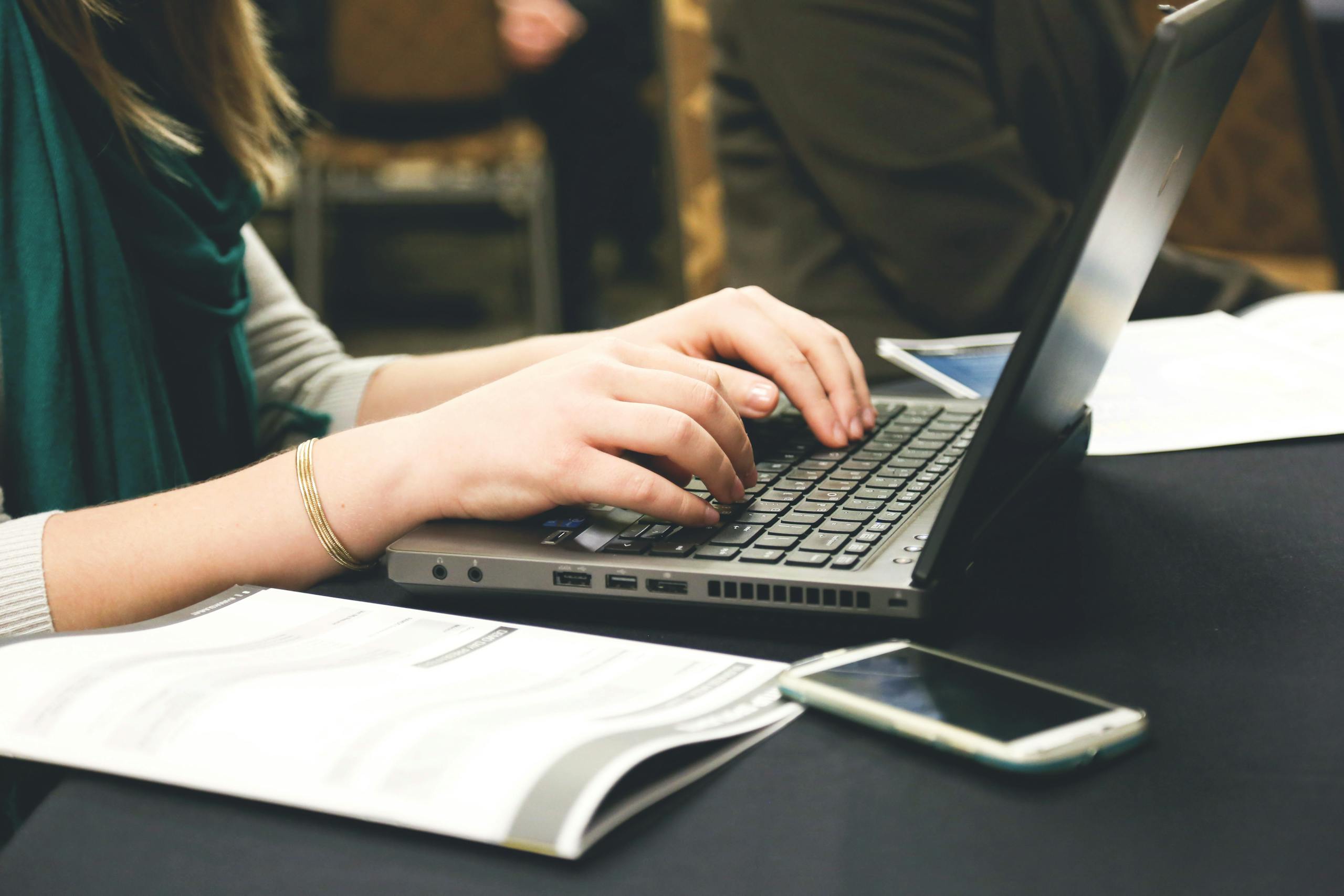 IPL 2026 Schedule PDF Download woman typing on a laptop at a desk with a smartphone and documents.