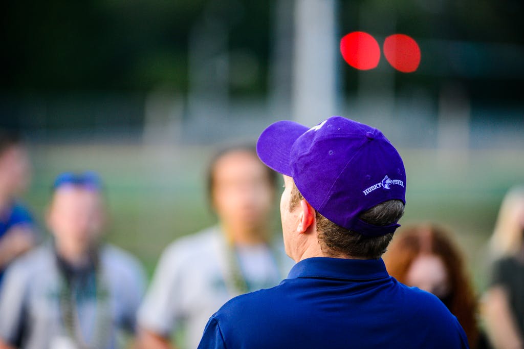 Purple Cap Standings A man in a purple cap observing a group outdoors during the day.