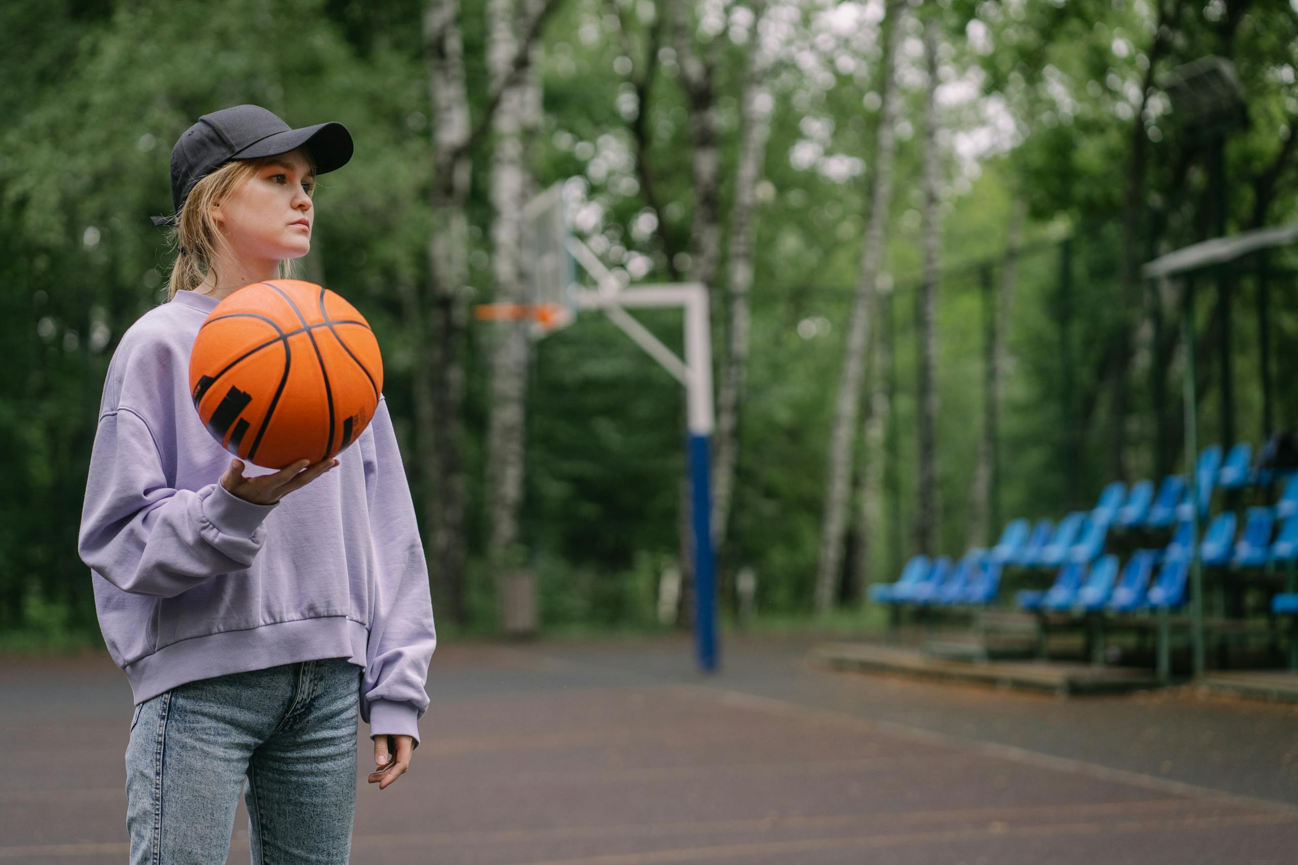 Purple Cap Standings Caucasian woman holding a basketball on an outdoor court, surrounded by trees.