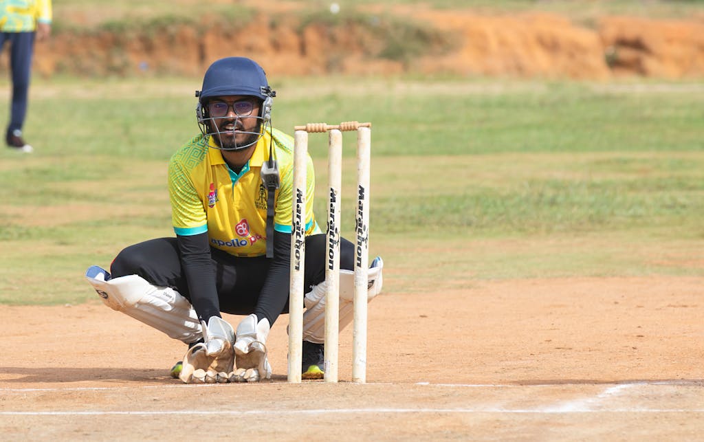 Dream11 Team Today Cricket wicketkeeper crouching behind stumps during a match on a sunny day on the field.