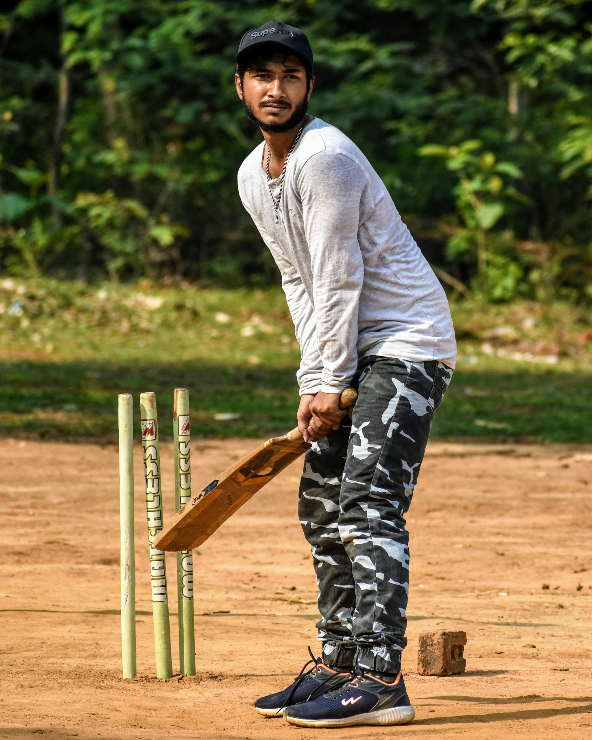 CSK New Captain Man holding a cricket bat in batting position on a rustic field with stumps.