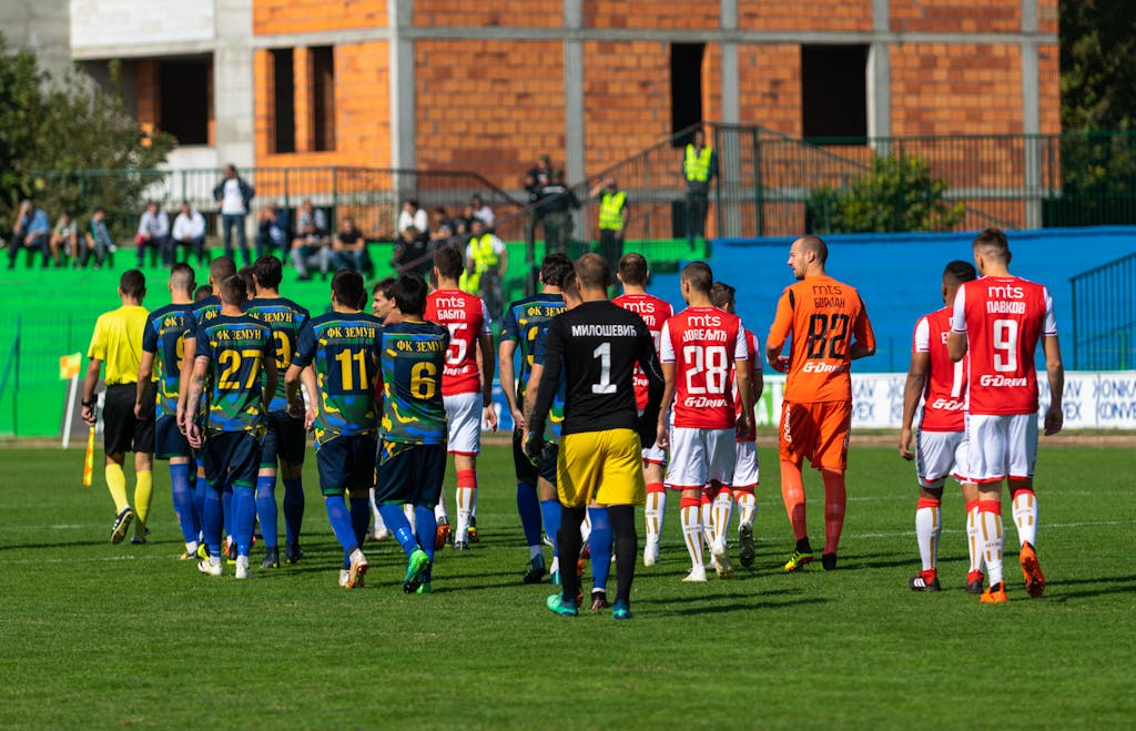 Dream11 Team Today Players from two teams in blue and red uniforms preparing for a football match on a green field.